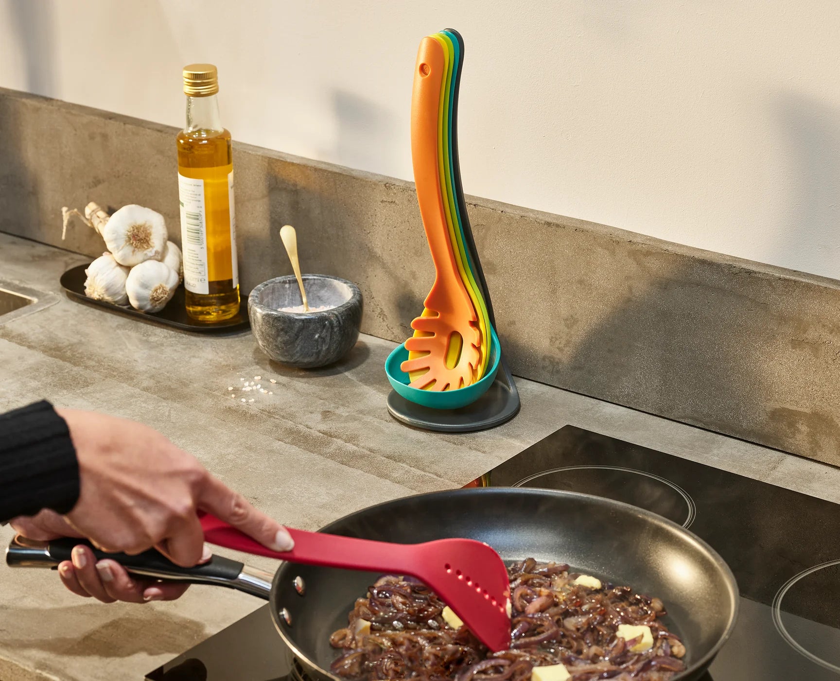 Red silicone utensil stirring food in a pan, with multicolour utensil on a teal stand nearby.