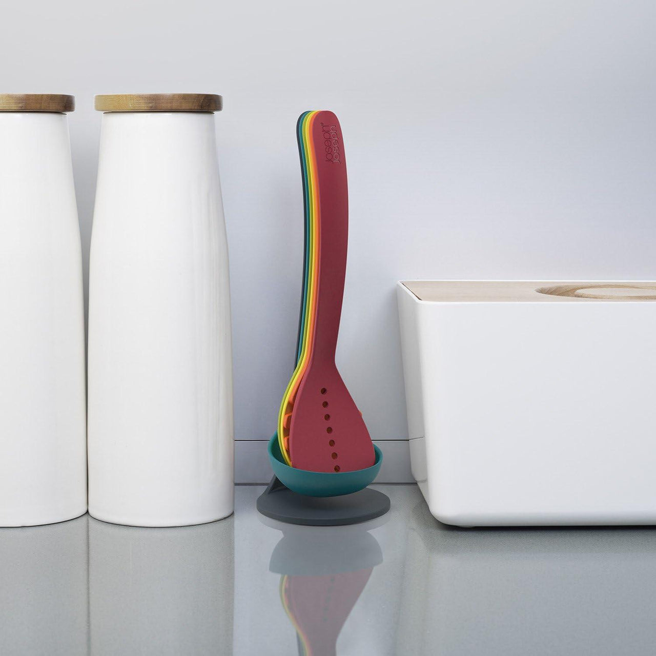 Single multicolour ladle utensil standing on teal magnetic base beside kitchen storage canisters.