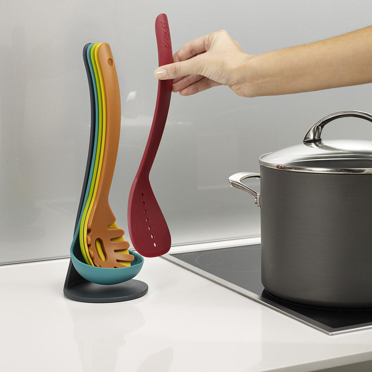 Hand placing a multicolour silicone utensil onto a teal magnetic stand next to a pot.