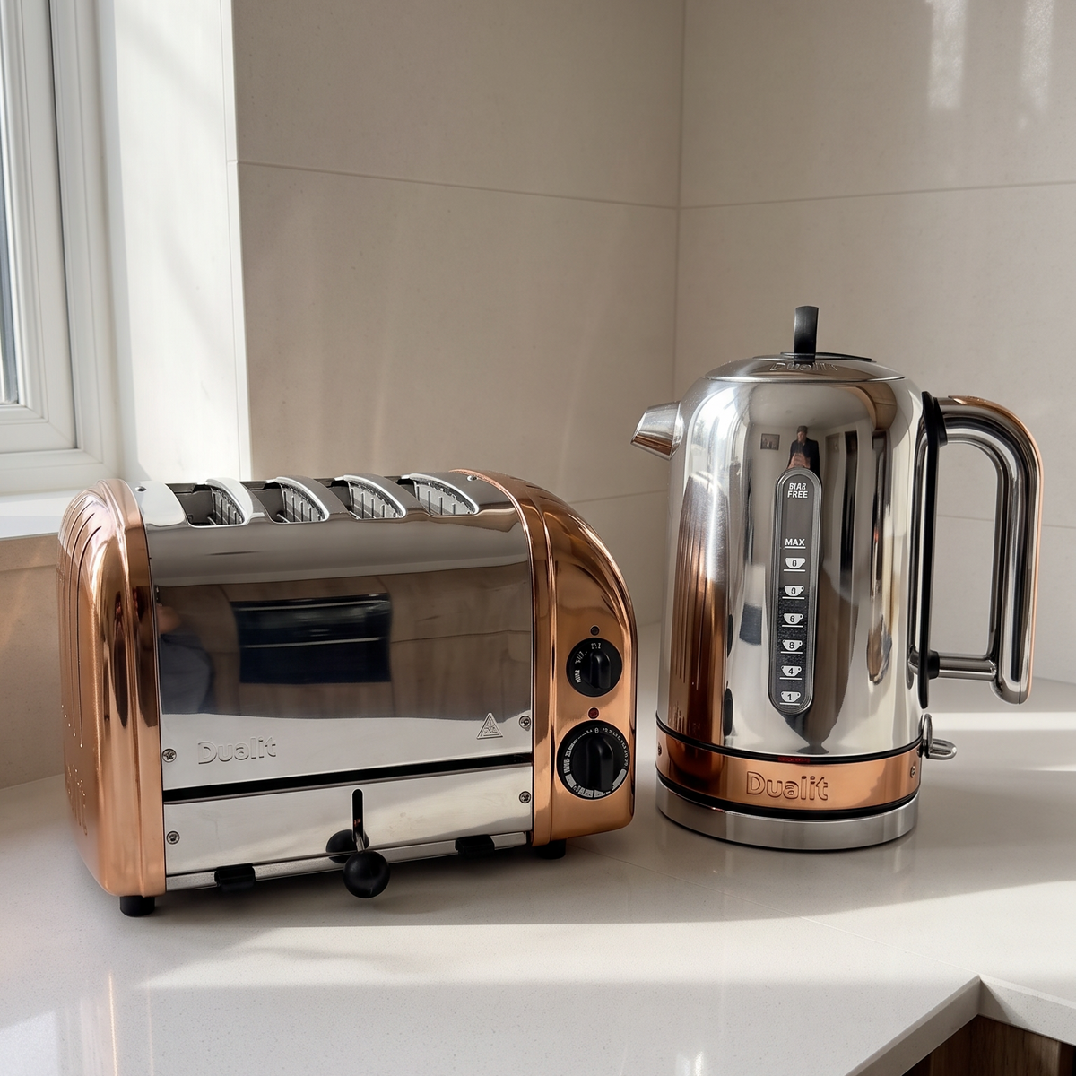 A copper and stainless steel 4-slice toaster sits on a white countertop next to a matching copper and stainless steel electric kettle in a modern kitchen setting.